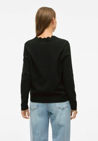 Zwarte gebreide trui met een geschulpte halslijn, lange mouwen en een aansluitende zoom, gedragen over lichtblauwe jeans. Glad textuur.