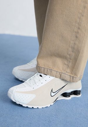 Personne portant des baskets Nike Shox beige avec un amorti noir, debout sur un tapis bleu, vêtue d'un pantalon beige.