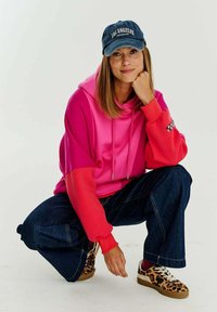 Hoodie s barevnými bloky v růžové a červené látce, s klokaní kapsou a šňůrkami. Nosený s tmavými džíny a teniskami s leopardím vzorem.