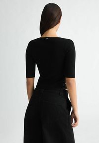 Schwarzes kurzärmeliges Strickoberteil mit Rundhalsausschnitt, versehen mit einem Knopfdetail auf der Rückseite. Kombiniert mit hochgeschnittenen schwarzen Jeans.