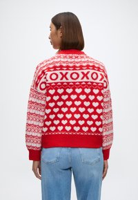 Suéter de punto rojo con patrones de corazones blancos y texto "XOXO". Tiene un cuello alto y puños de canalé, con un ajuste relajado y oversize.