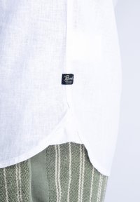 Gros plan de l'ourlet d'une chemise en lin blanc avec une petite étiquette noire indiquant « Petrol Industries Since 1989 » sur un tissu tricoté vert texturé.