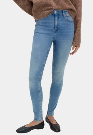 Lyseblå skinny jeans båret med en brun sweater og sorte ballerinaer på en stående person mod en ensfarvet baggrund.
