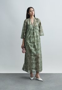 Γυναίκα με διαφανές πράσινο εμπριμέ μακρύ φόρεμα, κρατώντας μια μικρή ροζ τσάντα και φορώντας λευκές μυτερές γόβες, στέκεται μπροστά σε απλό φόντο.