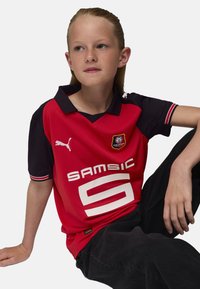 Camisa polo roja con mangas negras, que presenta un gran número blanco '5' y 'Samsic' impreso en negrita. Logo en el pecho. Fabricada en tejido ligero.
