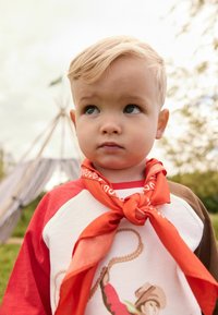Kind dat een wit shirt draagt met rode mouwen met een cartoonontwerp, en een geknoopte oranje bandana om de nek in een buitenomgeving.