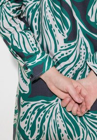 Camicia a maniche lunghe con un motivo di foglie verde e bianche su uno sfondo scuro, dotata di polsini con bottoni e tessuto morbido e strutturato.