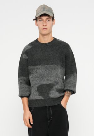 Junger Mann trägt einen grau-schwarz gemusterten Pullover, graue Mütze und schwarze Hose, mit der linken Hand in der Tasche vor einem einfarbigen Hintergrund.