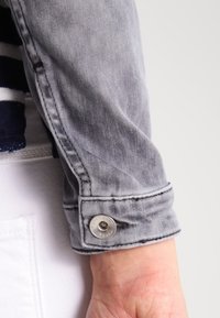 La foto ravvicinata del polso di una persona in una giacca di jeans grigia con un bottone metallico e un indumento bianco sotto.