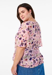Blusa in tessuto rosa con stampa floreale, maniche corte e svolazzanti. Presenta un orlo arricciato con fiori multicolori e foglie verdi.