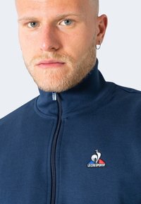 Chaqueta de cremallera en color azul marino, hecha de material suave, con un alto cuello y un logo con un emblema de gallo y acentos tricolores en el pecho.