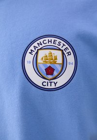 Šviesiai mėlynas džersis, kuriame yra apskritas logotipas su laivu, rože ir užrašu "Manchester City", akcentuotas auksinėmis ir mėlynomis detalėmis.