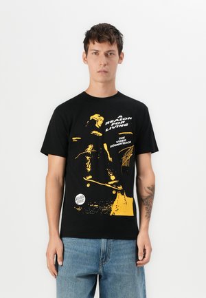 Zwart katoenen T-shirt met een gedurfde gele grafische afbeelding van een skateboarder, met tekst erboven en eronder, ronde hals, korte mouwen.