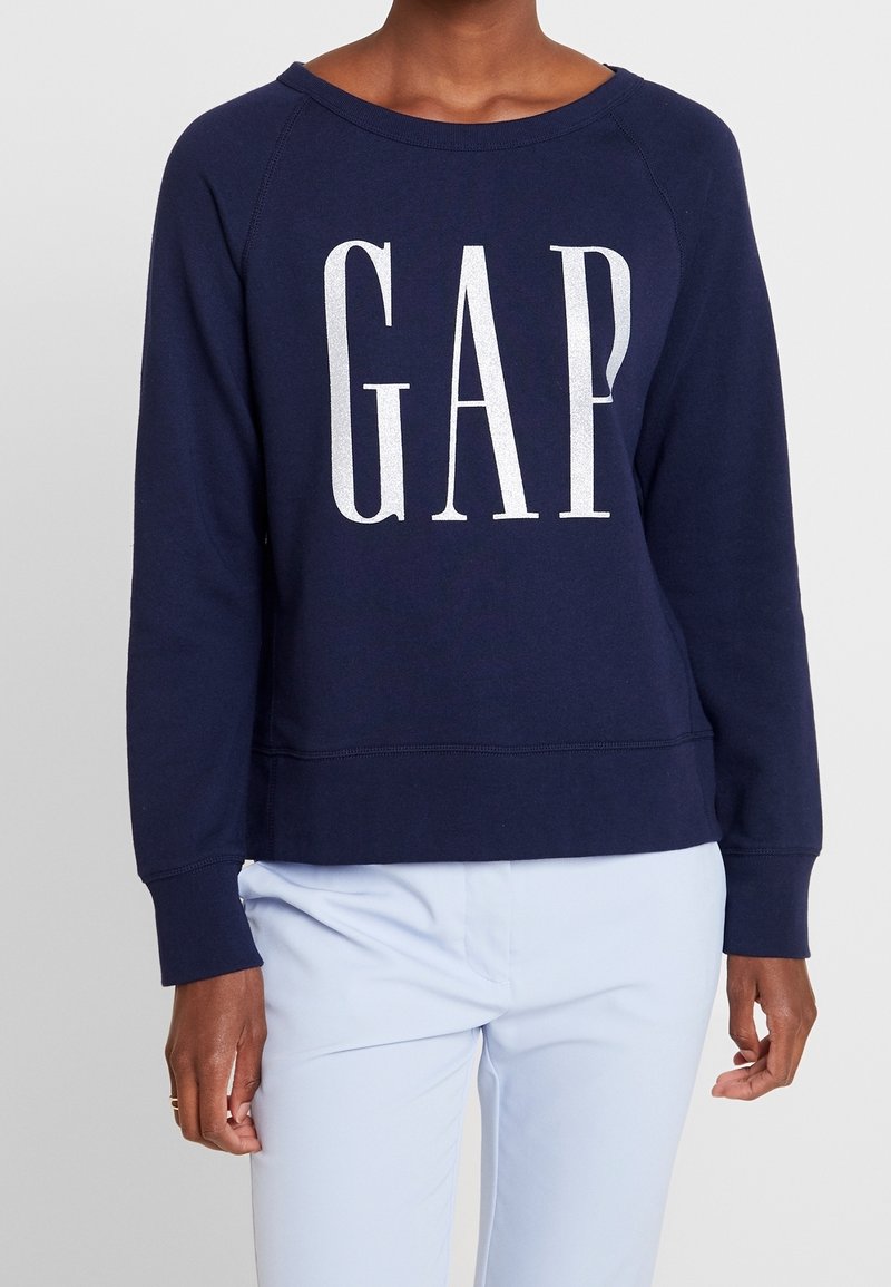 Sweat-shirt bleu marine avec grand logo blanc "GAP", porté avec un pantalon bleu clair, visible du cou jusqu'aux hanches.