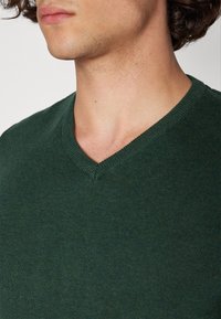 Pull vert à col en V fabriqué dans un tissu doux, présentant une texture en maille fine et un col côtelé, illustrant un gros plan de la zone du col.