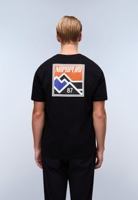 Juodas medvilninis marškinėlis su daugiamačiu grafikiniu dizainu, vaizduojančiu kalnus, ir užrašu "NAPAPIJRI 87" ant nugaros. Trumpomis rankovėmis, laisvas siluetas.