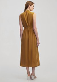Ärmelloses Midikleid in senfgelb, mit einem hohen Kragen und einem Taillengürtel. Sanfte Textur mit lässiger Passform, die unterhalb der Waden fällt.