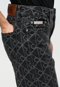 Σκούρο γκρι τζιν με σχέδιο δεμέν σε γεωμετρικά μοτίβα, ετικέτα δερμάτινης μέσης και ασημένιες μεταλλικές λεπτομέρειες.