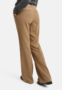 Wide-legged trousers in a tan fabric, featuring two back pockets with subtle stitching details, paired with a dark top and black shoes.