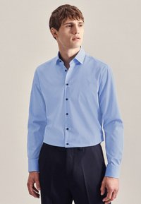 Camicia a maniche lunghe di colore azzurro chiaro con colletto classico, chiusura frontale con bottoni e bottoni neri. Realizzata in un tessuto liscio con una leggera trama.