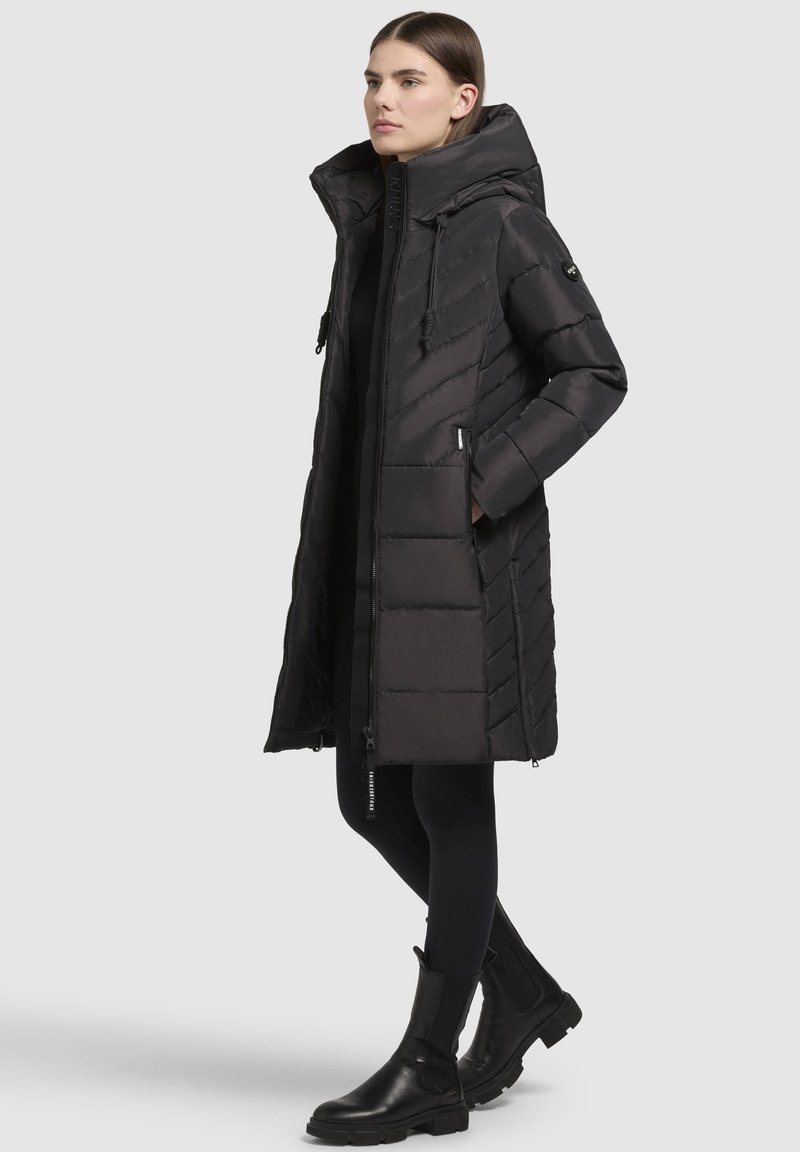 Schwarze Daunenjacke mit hohem Kragen, Reißverschluss, Seitentaschen und einem strukturierten Zickzackmuster. Getragen mit schwarzen Leggings und Stiefeln.