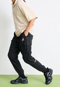 Person wearing beige shirt, black cargo pants with New Balance logo, and black sneakers, posing mid-step against white background and green floor.