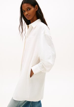 Kobieta o długich ciemnych włosach, ubrana w oversize'ową białą koszulę na guziki, z rękami w bocznych kieszeniach, zwrócona lekko bokiem na tle jednolitego tła.