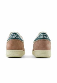 New Balancein tennarit, joissa on vaaleanpunainen mokkainen päällinen, valkoiset yksityiskohdat ja teal-väriä oleva kieli. Sisältää tekstuuriset pohjat ja brändilogiikan kantapäässä.