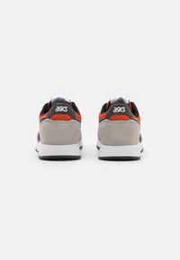 Sneakers grigi e neri con accenti rossi, dotati di un tallone in suede texturizzato e tomaia in rete, e una suola in gomma bianca. Logo visibile sul retro.