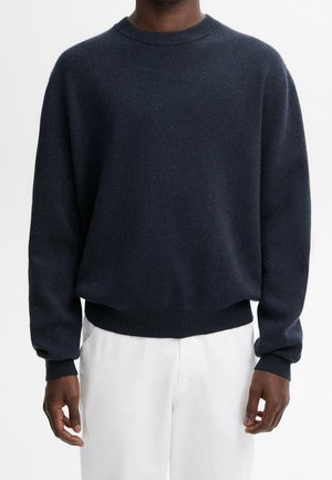 Pull en maille bleu marine avec col rond, poignets et ourlet côtelés. Texture douce avec de subtils flocons. Assorti à un pantalon blanc.