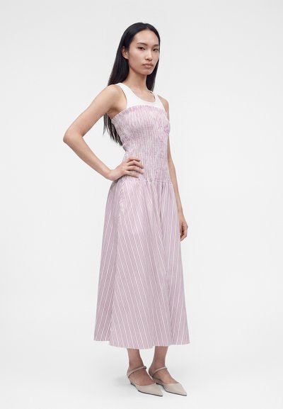 Γυναίκα με μακριά σκούρα μαλλιά φοράει αμάνικο midi φόρεμα με ροζ και λευκές ρίγες και μπεζ μυτερές μπαλαρίνες, στέκεται με το χέρι στη μέση.