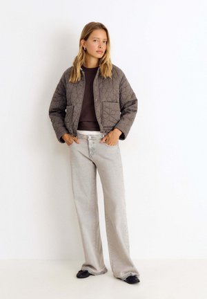 Joven mujer de pie con las manos en los bolsillos, vistiendo una chaqueta gris acolchada, camisa oscura, vaqueros claros de pierna ancha y zapatos negros sobre fondo blanco.