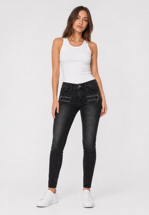 Kvinde stående iført hvid tanktop, sorte skinny jeans med lynlåse og hvide sneakers mod en ensfarvet lys baggrund.
