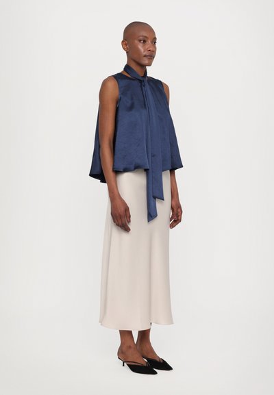 Navy satin sleeveless top with a neck tie, paired with a light beige, ankle-length skirt and black pointed slip-on shoes.