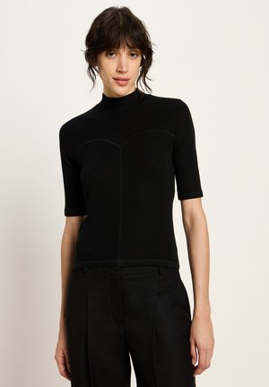 Woman wearing a black fitted short-sleeve top with high neck and black tailored pants, standing against a plain light background.