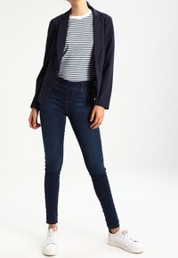 Námořnický blazer přes bílý a námořnický pruhovaný tričko, spárovaný s tmavými skinny džíny a bílými teniskami. Oblečení je ležérní a přiléhavé.