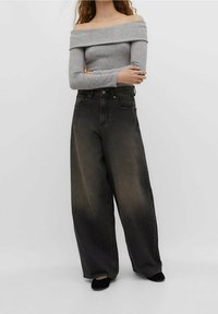 Femme portant un pull gris côtelé à épaules dénudées, un jean noir délavé à taille haute et large, et des chaussures noires, debout les bras croisés.