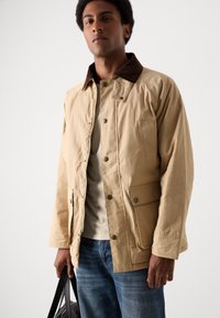 Beige jakke med en brun kordfløyelskrage, trykknapper, to frontlommer og glidelås, båret over en lys skjorte og blå jeans.