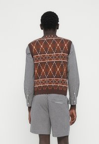 Άνδρας που φοράει καφέ πλεκτό γιλέκο με σχέδια πάνω από ασπρόμαυρο καρό πουκάμισο και ταιριαστά καρό σορτς, φαίνεται από πίσω μπροστά σε άσπρο τοίχο.