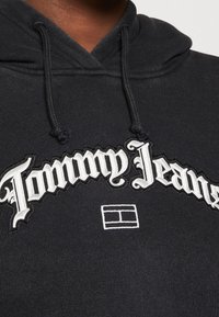 Czarna bluza z kapturem z białym haftowanym logo "Tommy Jeans" i prostokątnym emblematem. Materiał wydaje się miękki, wykończony teksturowanym detalem.