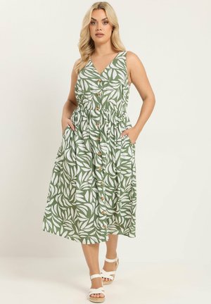 Blondynka ubrana w bezrękawnikową zieloną sukienkę midi z białymi wzorami liści, zapinaną na guziki z przodu, z bocznymi kieszeniami oraz białe sandały na otwarte palce.