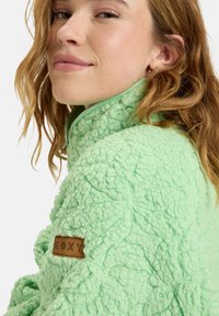Green fleece jacket with a textured, fluffy surface. Features a high collar and a brown leather logo patch on the left shoulder.