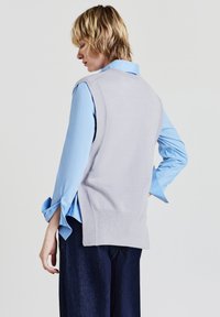 Pullover senza maniche grigio chiaro con orlo a coste indossato sopra una camicia a maniche lunghe blu chiaro. La camicia ha polsini arrotolati e una vestibilità comoda.