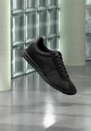 Pantof sport negru cu talpă texturată și șireturi, suspendat deasupra unei podele reflectorizante de beton lângă un perete din blocuri de sticlă și o coloană mare.