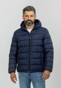 Tamsiai mėlynas pūstas džemperis su kapišonu, turintis užtrauktuką priekyje, horizontalius siūlus ir šonines kišenes. Dėvimas ant baltos marškinių ir džinsų.