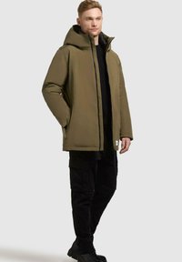 Olivgrüne isolierte Jacke mit Kapuze, vorderem Reißverschluss, Seitentaschen und schwarzer Innenauskleidung. Kombiniert mit schwarzen Cargo-Hosen und Stiefeln.