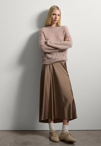 Donna con lungo capelli biondi, che indossa un maglione di maglia beige, una gonna midi di raso bronzeo, calzettoni bianchi e sneakers beige, in piedi con le braccia incrociate.