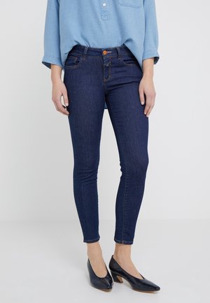 Jeans ajustados de color azul oscuro con una textura suave, de cintura alta y costuras naranjas. Combinados con una camiseta de botones de color azul claro y zapatos de tacón negros.