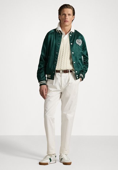 Chaqueta bomber de satén verde con rayas blancas, suéter blanco de punto trenzado, pantalones blancos y zapatillas verdes de acento. Texturas suaves y diseño ajustado.