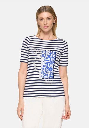 Femme portant un t-shirt à manches courtes rayé bleu marine et blanc avec un écusson floral bleu et un texte sur la poitrine, assorti à un pantalon blanc.